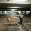 A solitary man leans on a shopping cart in an empty parking garage, evoking a sense of loneliness and urban struggle.