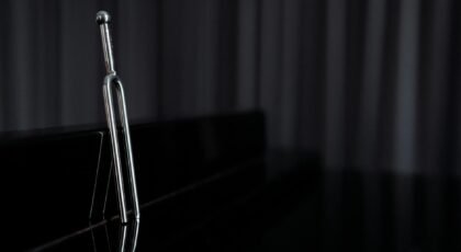 A close-up of a metal tuning fork standing on a reflective piano surface with dark background.