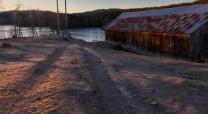 Abandoned barn next to a frozen lake with a sunset backdrop, creating a serene winter scene.