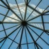 Captured from below, a geometric glass rooftop showcases the clear blue sky above.