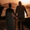 A couple walking hand in hand at sunset on a scenic coastal path, silhouetted against the sea.