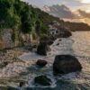 Scenic view of Bali coastline with rock formations and lush greenery at sunrise.