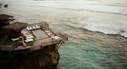 Aerial view of a luxurious cliffside resort in Bali, Indonesia, with ocean and swimmers below.