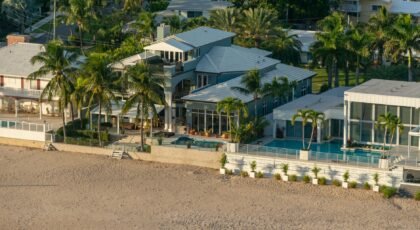 Aerial view of luxury beachfront houses with palm trees, pool, and sandy beach.
