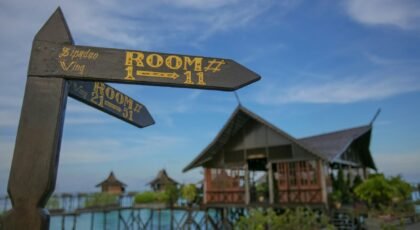Wooden signage directing rooms at a tropical beach resort with clear blue skies and stilt houses.