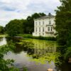 A tranquil scene of a white mansion by a lily-covered pond surrounded by lush greenery.