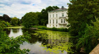 A tranquil scene of a white mansion by a lily-covered pond surrounded by lush greenery.