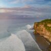 Breathtaking aerial view of Uluwatu cliffs in Bali with vibrant ocean waves during sunset.