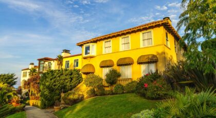 Sunny view of a Mediterranean-style villa surrounded by lush greenery and blue sky.
