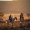 Two riders enjoy horseback riding in a valley surrounded by hot air balloons at sunset.