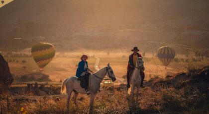 Two riders enjoy horseback riding in a valley surrounded by hot air balloons at sunset.